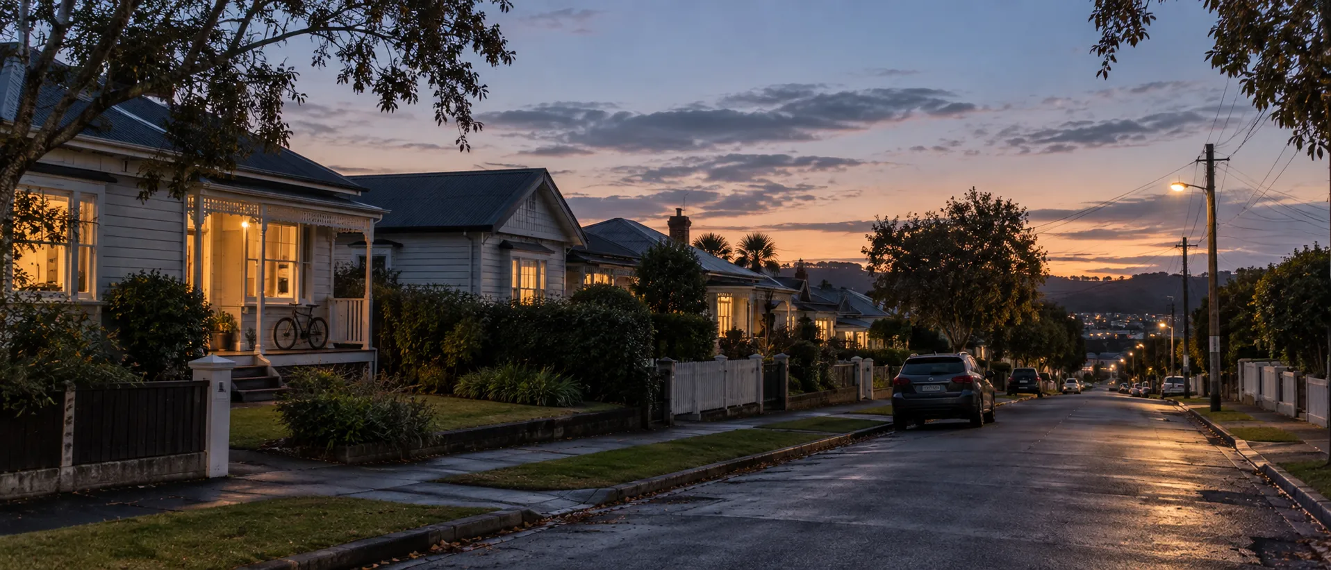 A suburban New Zealand street at dusk with lights on in the houses